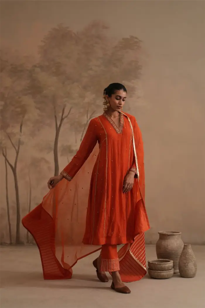 Model wearing a rust-orange embroidered kurta set with sheer dupatta